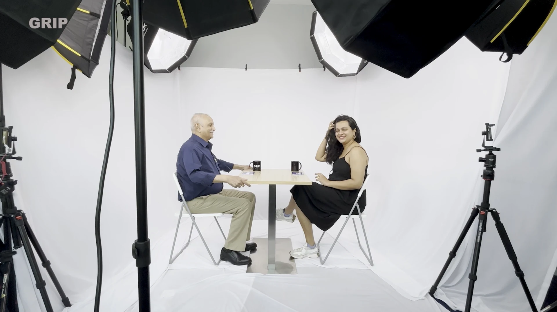Two people seated at a table in a white studio surrounded by lights and cameras.